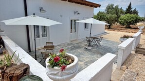 A white building with two large white umbrellas, a patio with chairs, and a potted plant with red flowers.