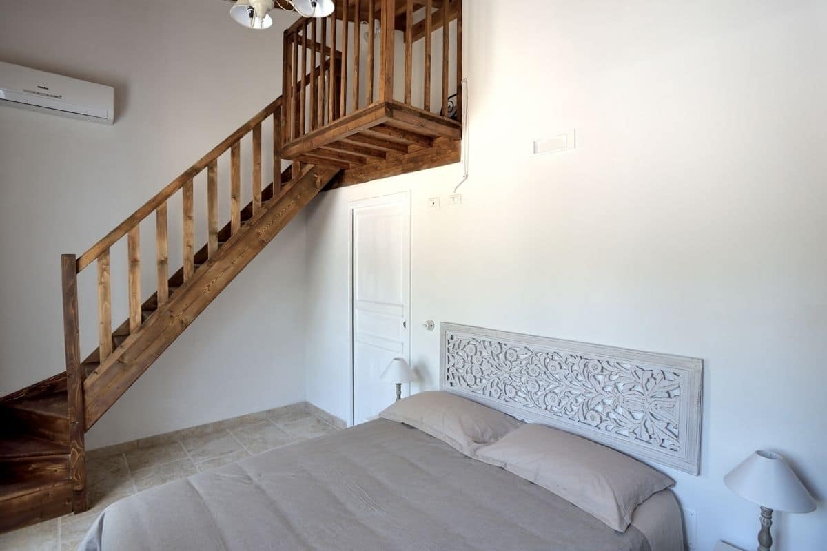 Une chambre avec un escalier en bois, un lit doté d’une tête de lit décorative et un mur blanc sur lequel est installé un appareil de climatisation.