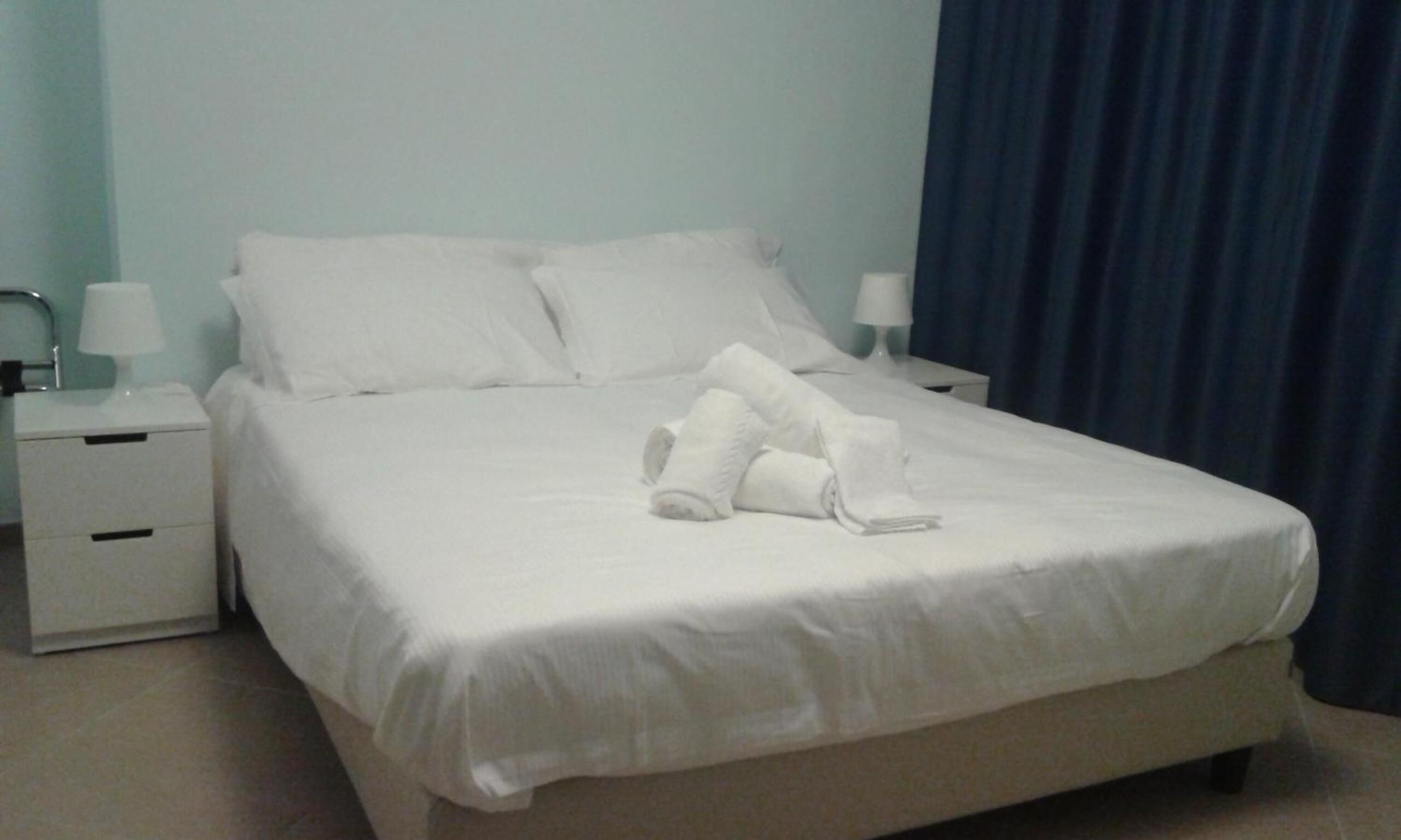 Cama bem arrumada com lençóis e toalhas brancos, uma mesa de cabeceira com abajur e um criado-mudo com abajur.