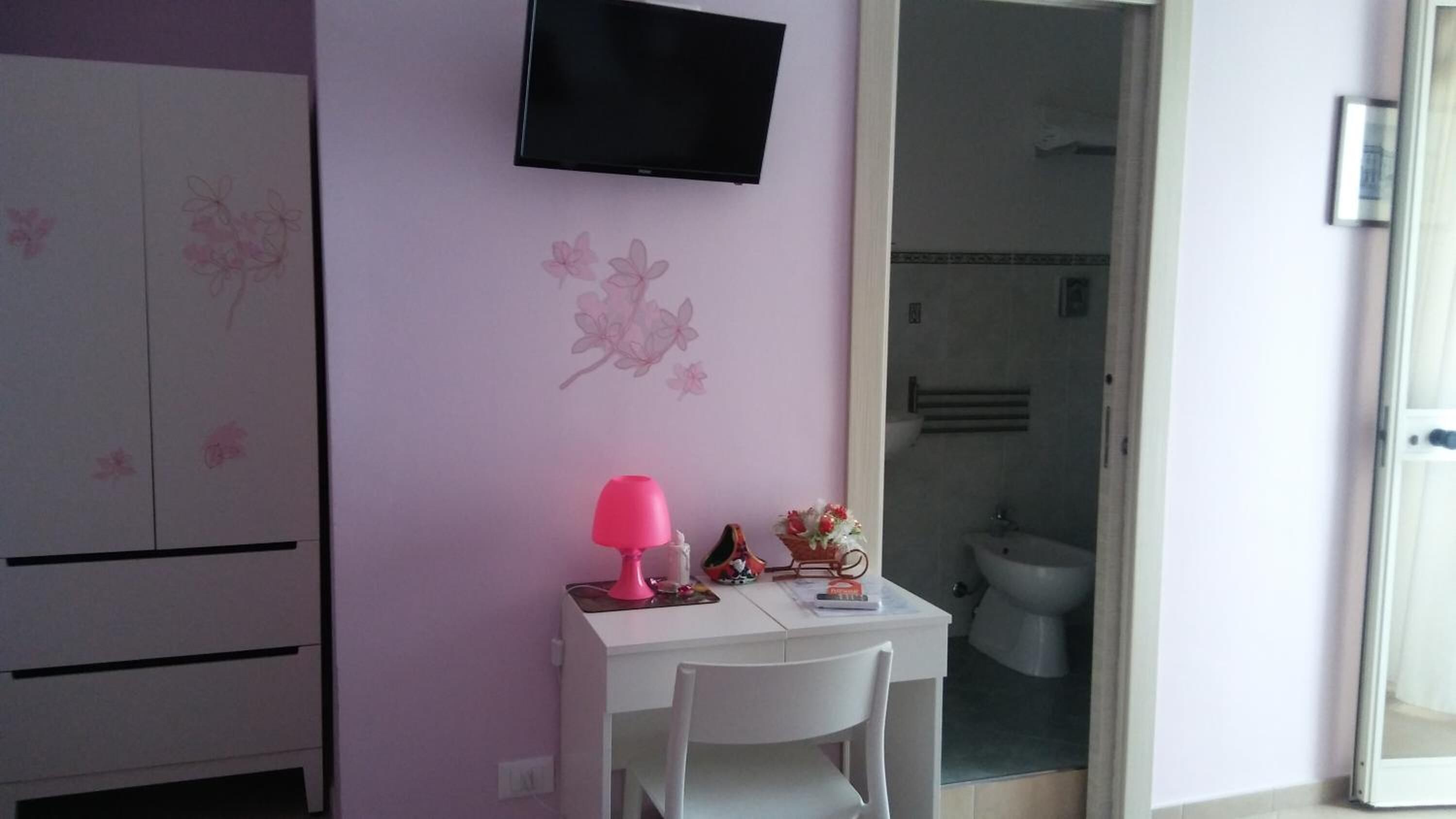 A small room with a TV mounted on the wall, a desk with a pink lamp, and a chair. There is a wardrobe with floral designs and a bathroom visible through an open door.