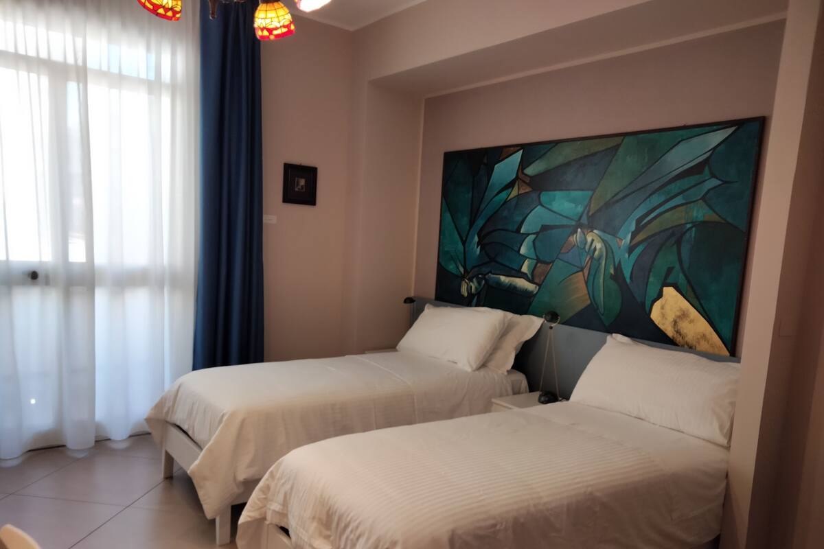 Habitación de hotel con dos camas, un cuadro abstracto grande en la pared y una ventana con cortinas translúcidas.