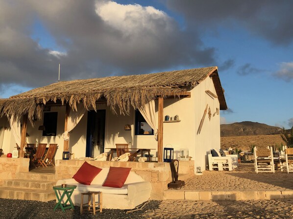 Un bâtiment rustique avec un toit de chaume, une véranda avec des chaises en bois et un canapé blanc agrémenté de coussins rouges.