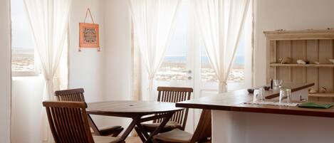 Un coin repas avec une table et des chaises en bois, une petite étagère ornée d’objets décoratifs et une vue sur la plage à travers de fines rideaux.