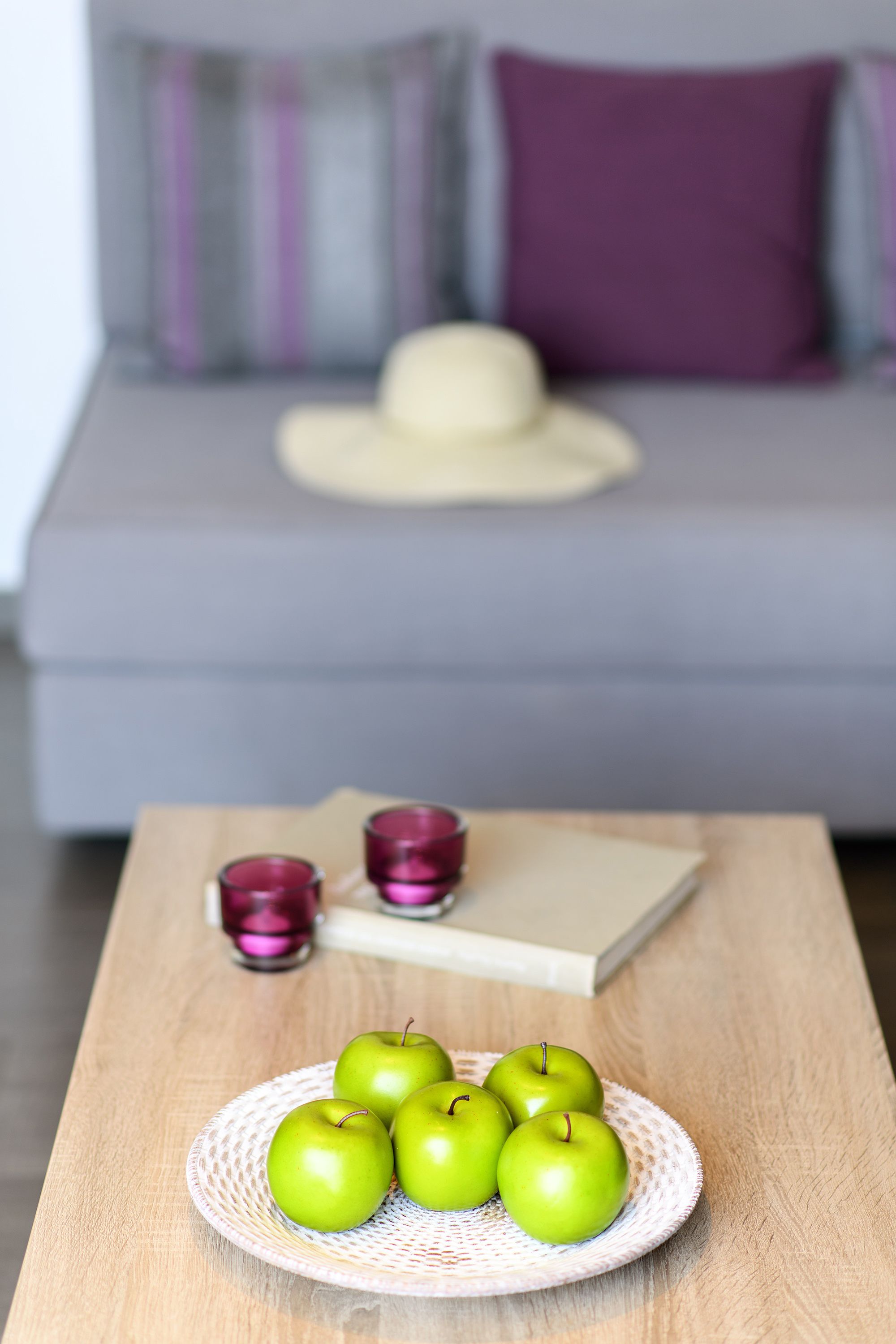 Une table en bois sur laquelle se trouve une assiette blanche, avec quatre pommes vertes, un livre et deux bougies violettes.