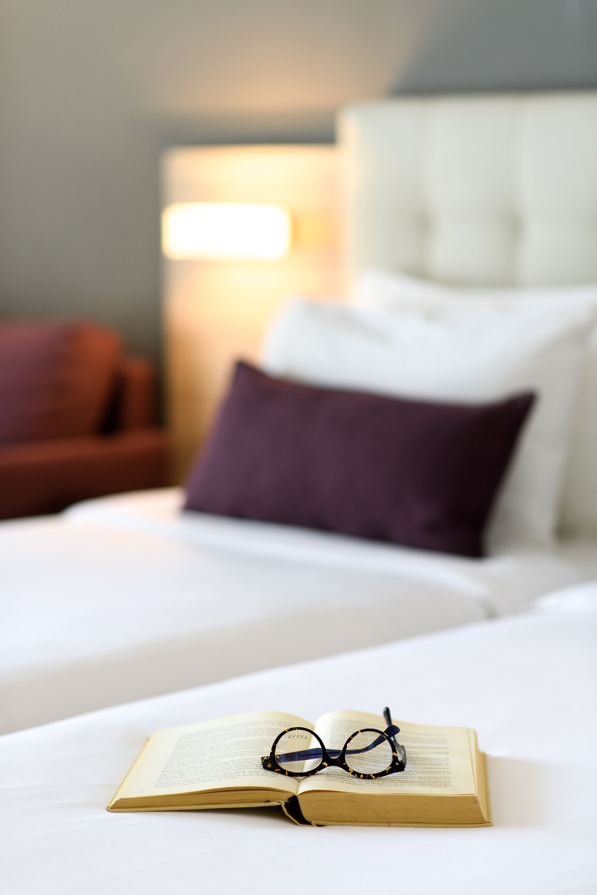 A neatly made bed with a book and glasses on it, a purple pillow, and a red chair in the background.
