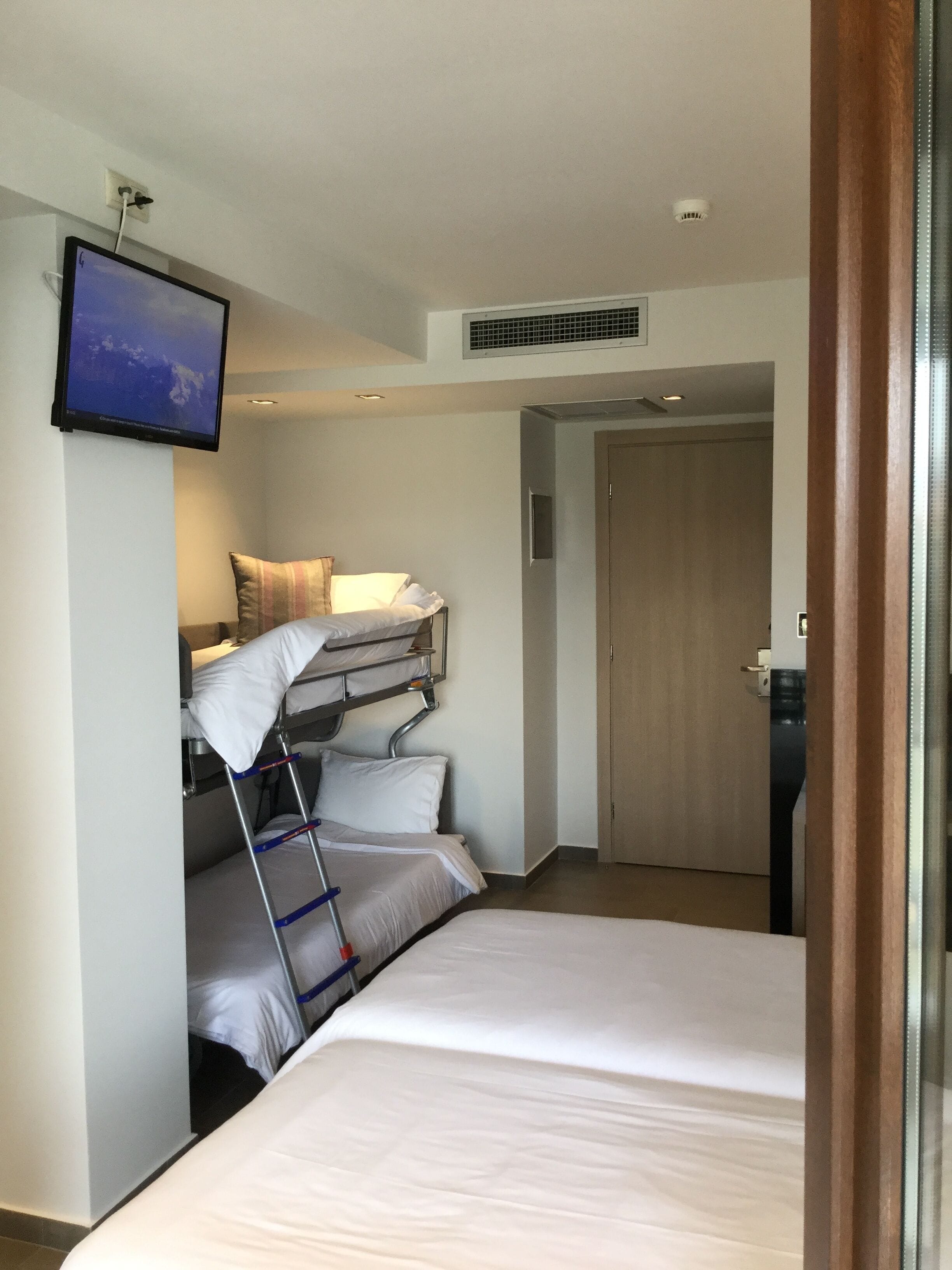 A hotel room with a bunk bed, a flat bed, a television mounted on the wall, and a door leading to another room.