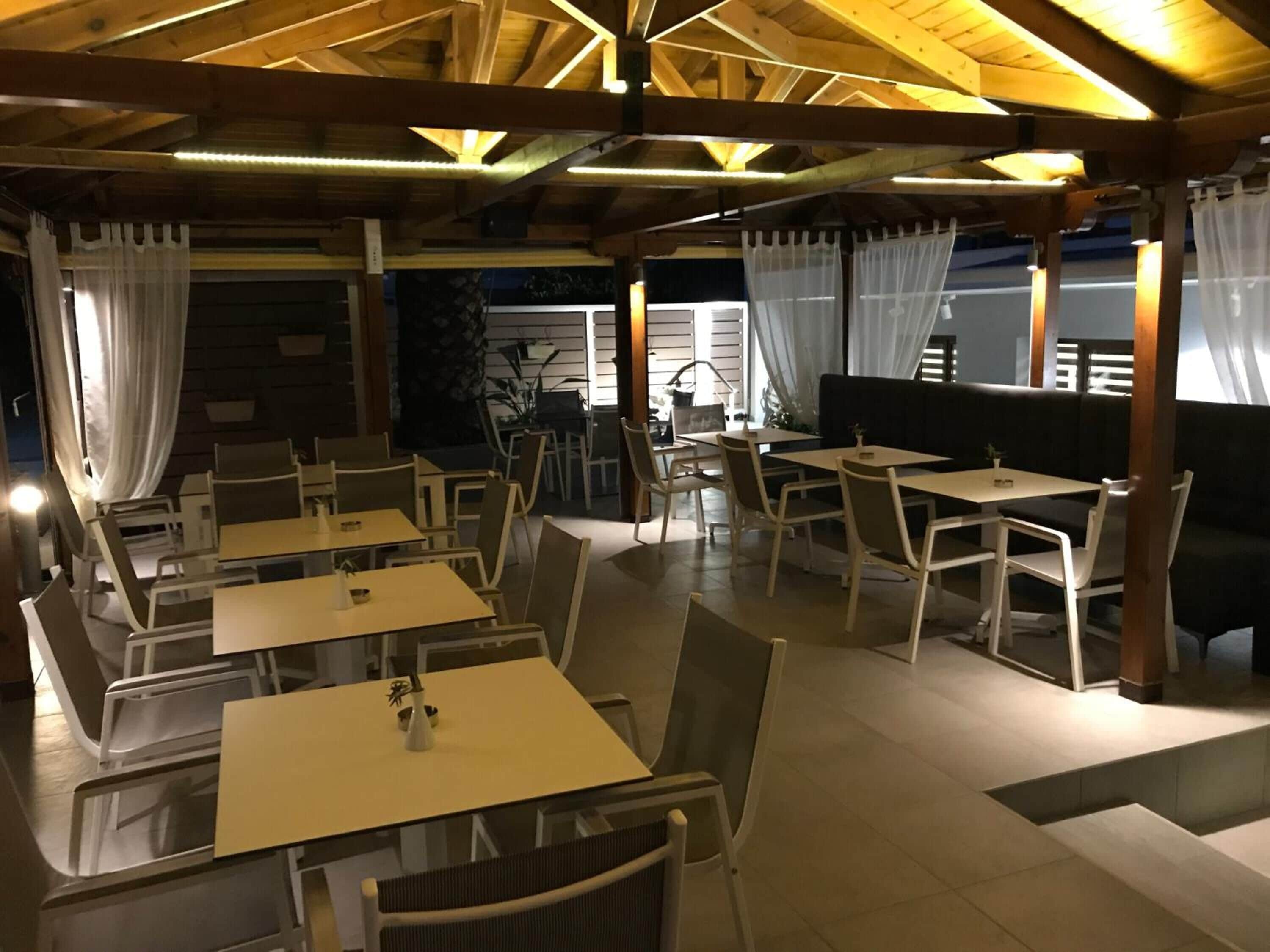 Un restaurante al aire libre con varias mesas y sillas, vigas de madera en el techo y cortinas blancas.