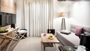 Quarto de hotel moderno com escrivaninha de madeira, sofá, mesa de jantar com frutas e bebidas, luminária e TV.