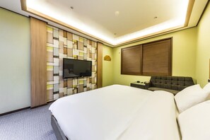 Ein modernes Hotelzimmer mit einem großen Bett, einem Flachbildfernseher an einer Wand mit geometrischem Muster, einem Holzschrank, einem kleinen Tisch mit einer Lampe und einem Fenster mit Jalousien.