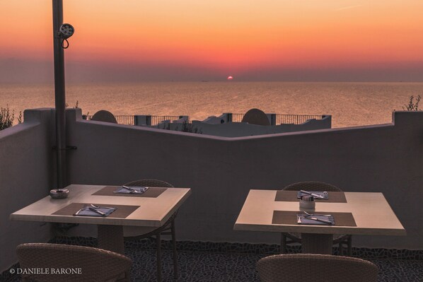 Una terraza con dos mesas dispuestas para comer, con vista a la puesta de sol sobre el mar.