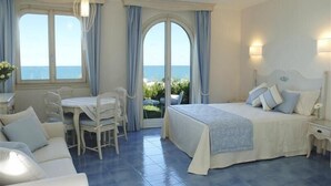 Un dormitorio con cama, escritorio y silla. Hay dos ventanas con cortinas, y se ve una vista al mar a través de ellas.