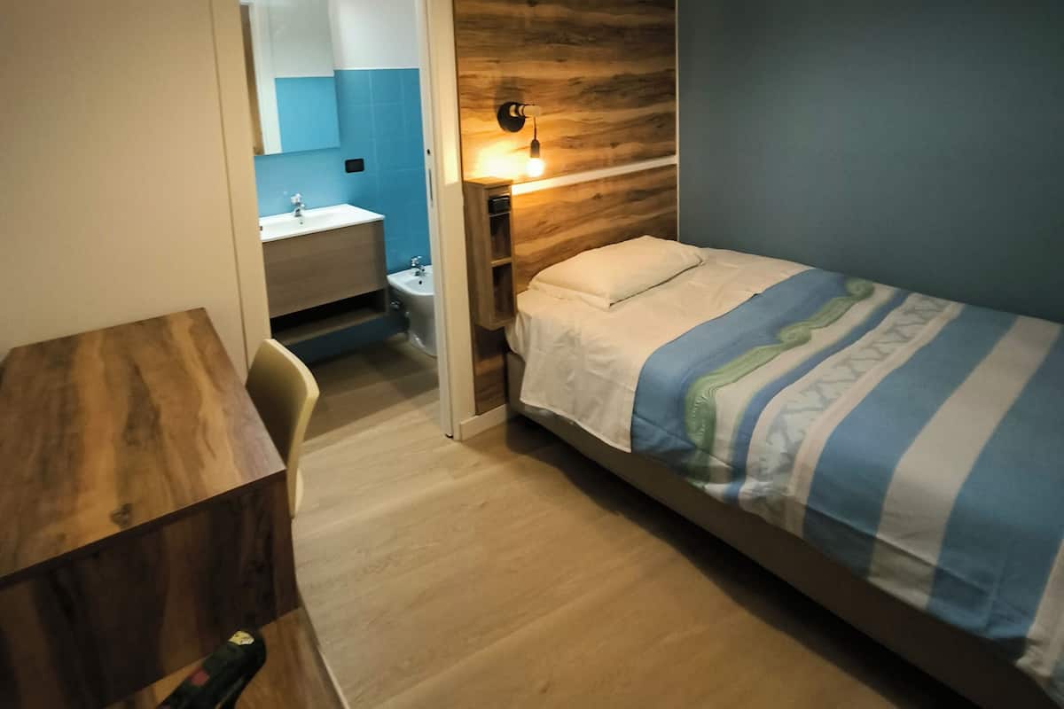 A bedroom with a bed, bedside table, and a nightstand with a lamp. There is a bathroom visible through an open door.