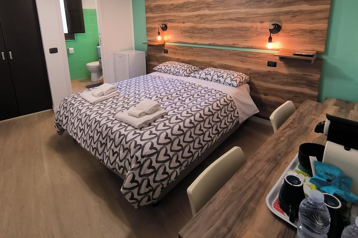 A modern hotel room with a wooden headboard, a bed with patterned bedding, a bedside table with a tray of toiletries, and a view into the bathroom.