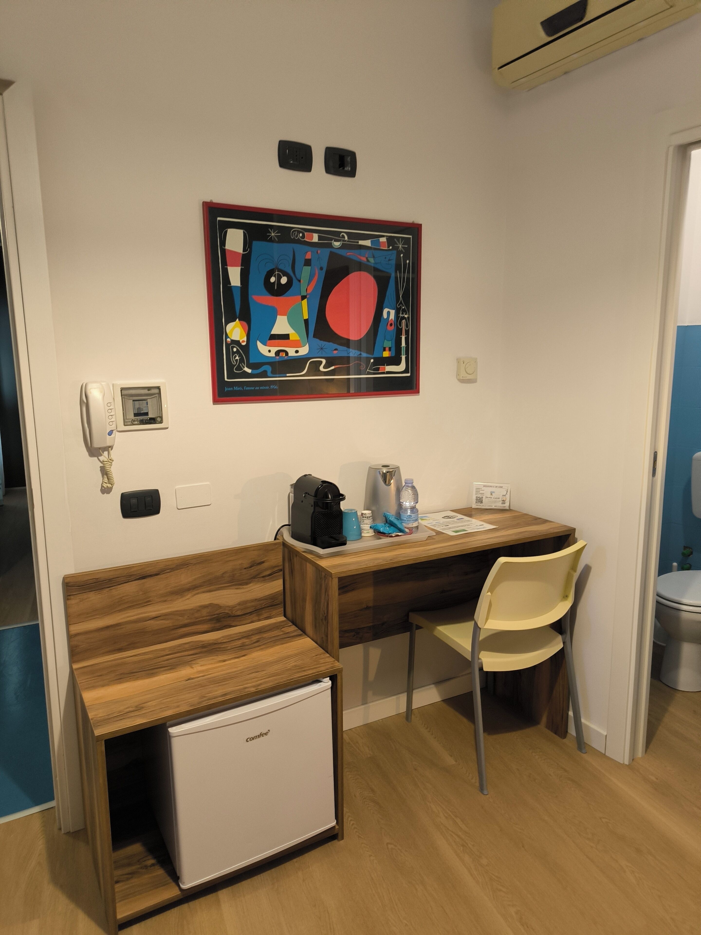 Een compacte kamer met een houten bureau, een kleine koelkast, een stoel, een koffiezetapparaat en een aan de muur bevestigd telefoon.