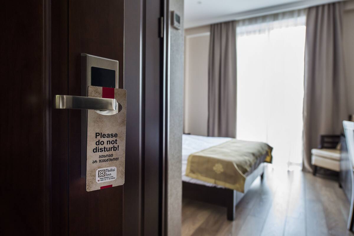 Une chambre d’hôtel avec un lit, un bureau et une fenêtre avec des rideaux. Il y a une pancarte sur la porte qui indique : « Veuillez ne pas déranger. »