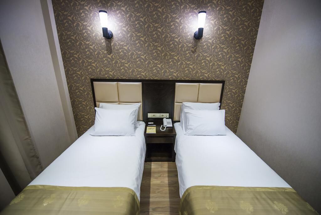 Camera d'albergo con due letti singoli, pavimento in legno, carta da parati decorata e due luci fissate al muro.