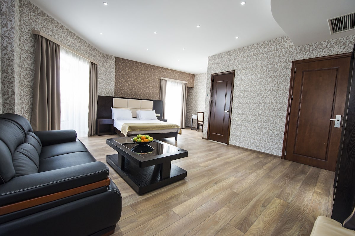 Une chambre d’hôtel moderne avec un grand lit, un canapé en cuir noir, une table basse avec des fruits et un plancher en bois.