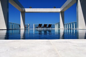 Area piscina moderna con cielo azzurro limpido, struttura geometrica a bordo vasca e lettini prendisole.