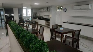 A modern dining area with a long table and chairs, a kitchen area with cabinets and appliances, and a clock on the wall. - Val Atlantic Hotel (João Pessoa)