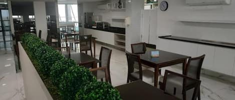 Un comedor moderno con una mesa larga y sillas, una zona de cocina con armarios y electrodomésticos, y un reloj en la pared.