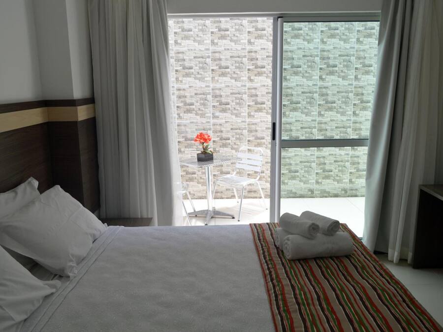 A hotel room with a bed, a small table with a flower, and a view of a brick wall through a sliding door.