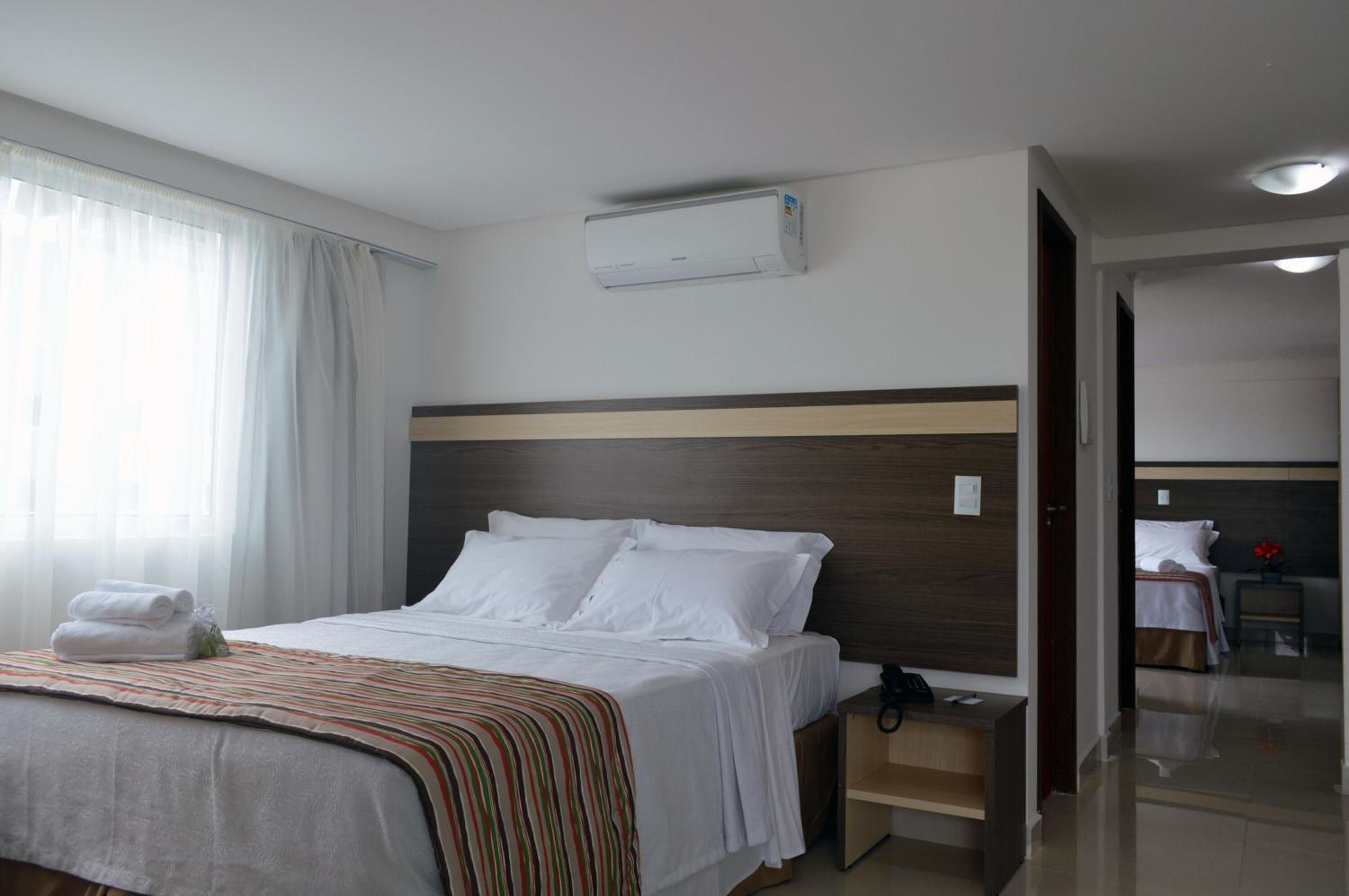 Una habitación de hotel con cama, mesita de noche, unidad de aire acondicionado y vista a otra habitación.