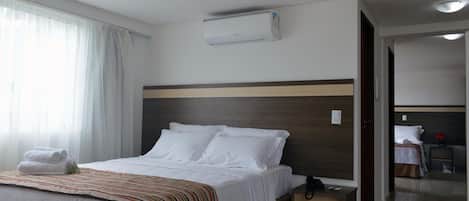 Una habitación de hotel con cama, mesita de noche, unidad de aire acondicionado y vista a otra habitación.
