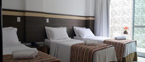 Habitación de hotel con tres camas, un ventanal con vistas y una mesita con un jarrón de flores.