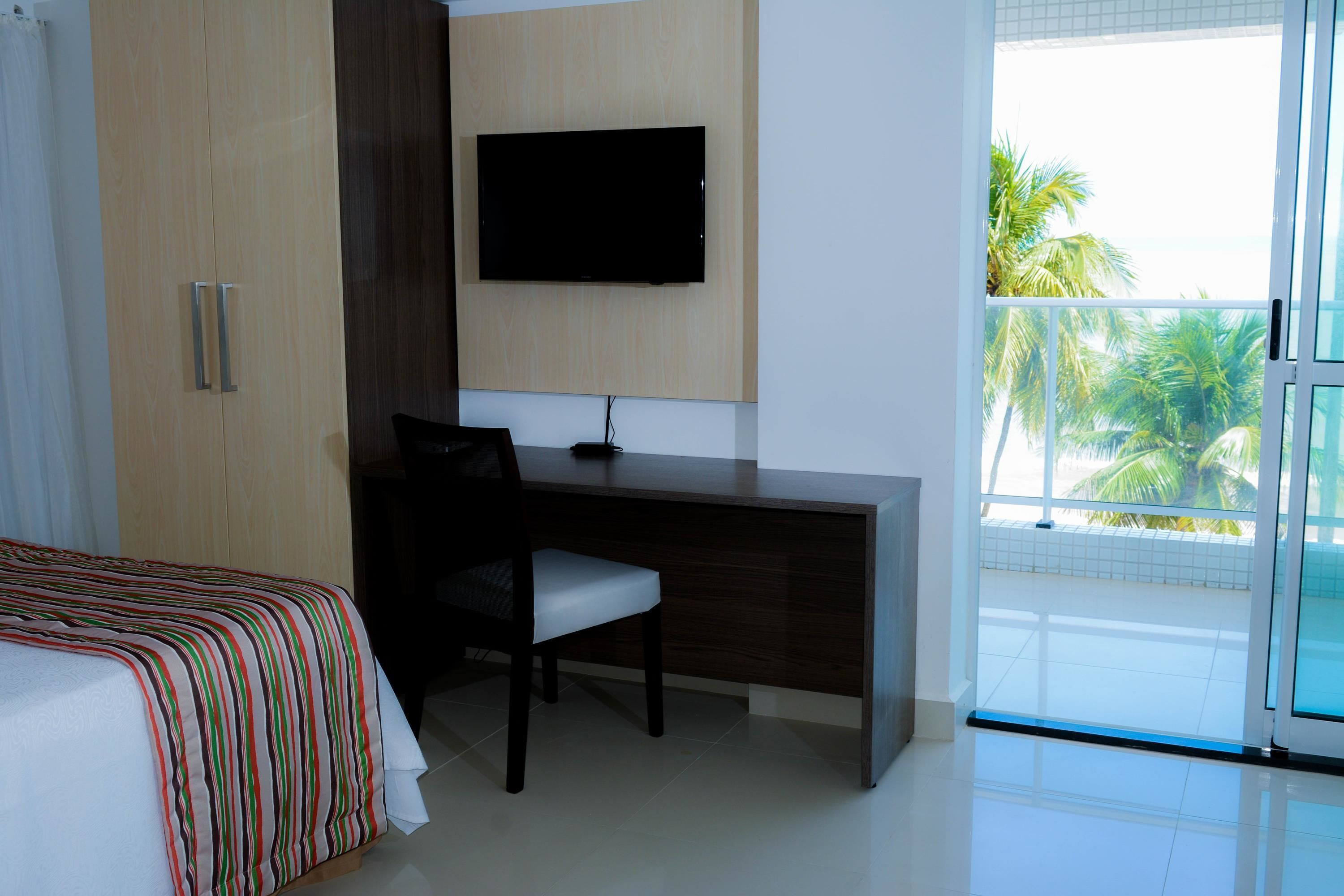 A hotel room with a bed, a desk, a chair, a TV mounted on the wall, and a balcony with a view of palm trees.