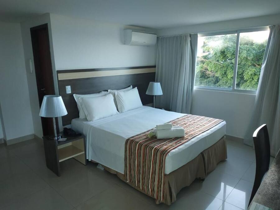 A hotel room with a large bed, two bedside tables with lamps, an air conditioning unit, a window with curtains, and a view of greenery outside.