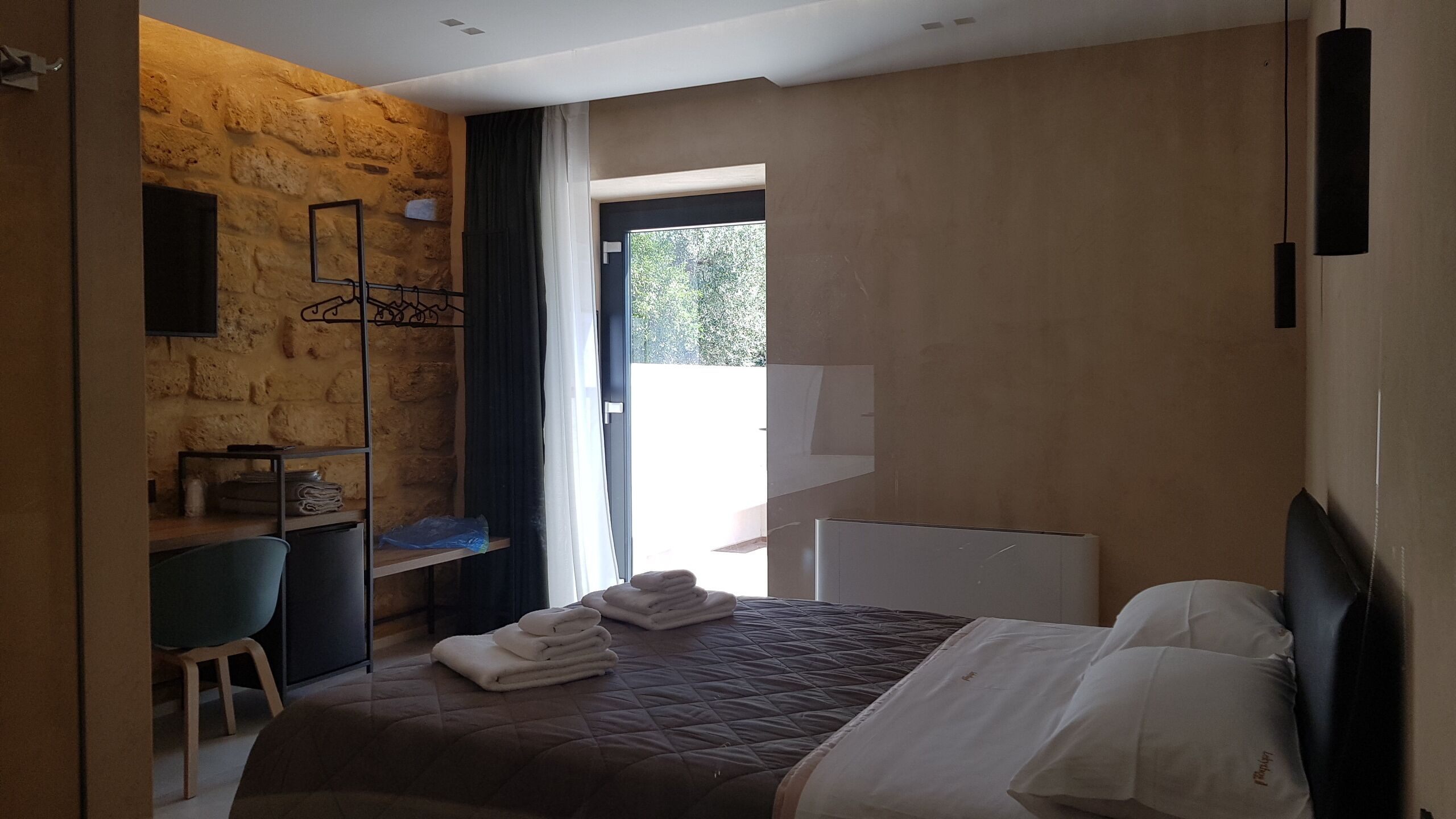 Ein modernes Schlafzimmer mit einer Wand aus Stein, einem Bett mit weißen Leinen, einem Schreibtisch mit Stuhl und Blick ins Grüne.