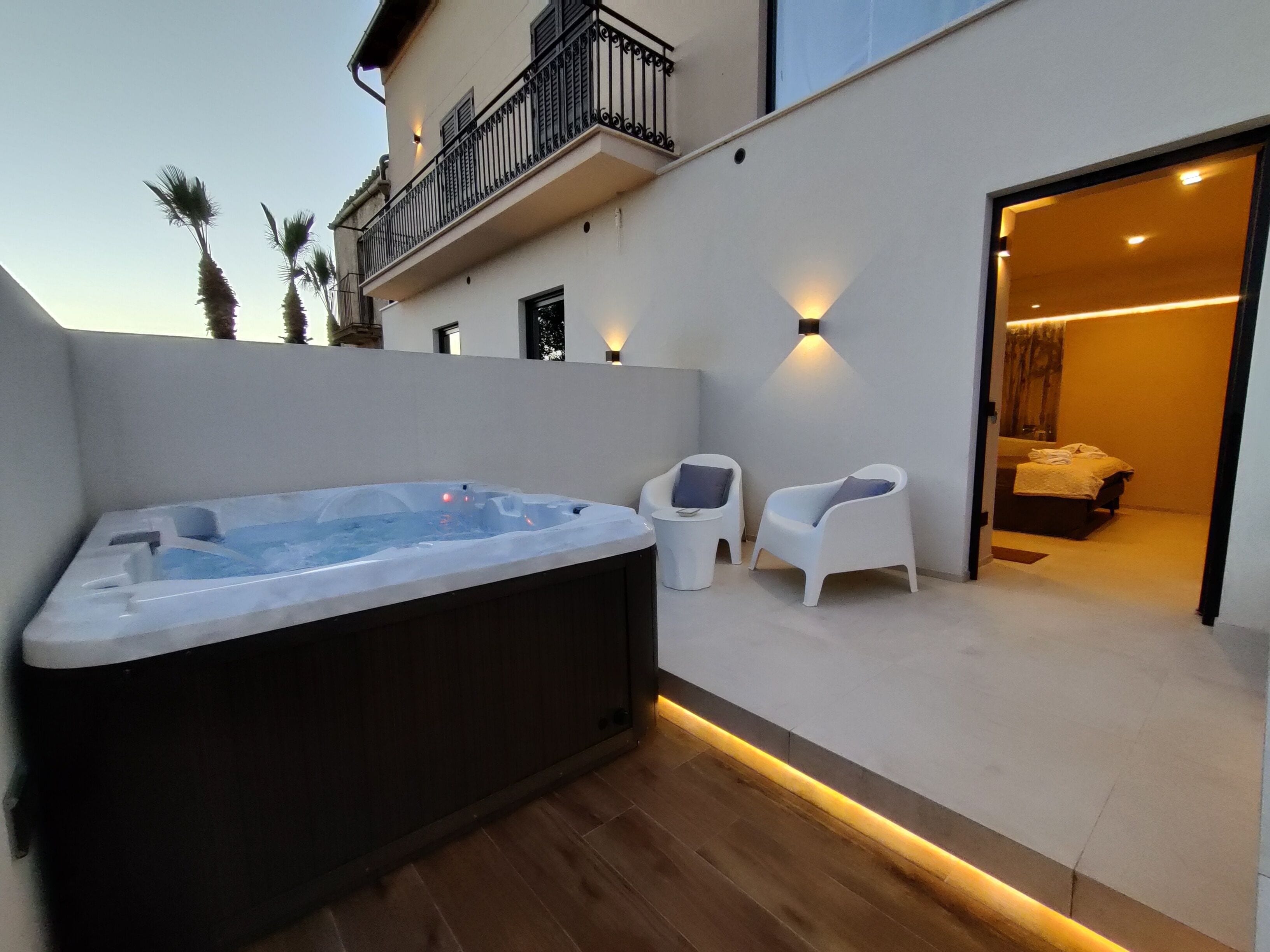 Un espacio exterior moderno con un jacuzzi, dos sillones blancos y una habitación visible a través de una puerta abierta.