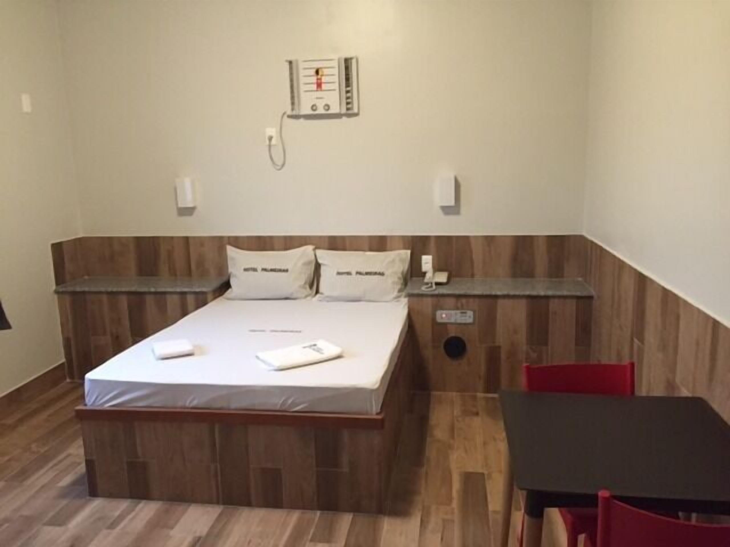 Habitación de hotel con cabecera de madera, cama con ropa blanca, mesita de noche, una silla roja y una pequeña mesa con mantel negro.