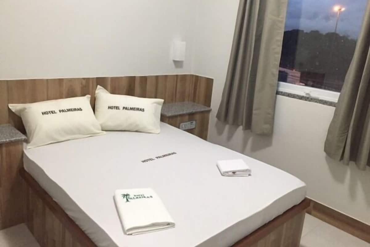 A hotel room with a bed, two pillows with the hotel's name, a nightstand, a window with curtains, and a folded towel on the bed.