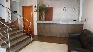 Una zona de recepción con mostrador, escalera, puerta de madera, planta y sofá de cuero.