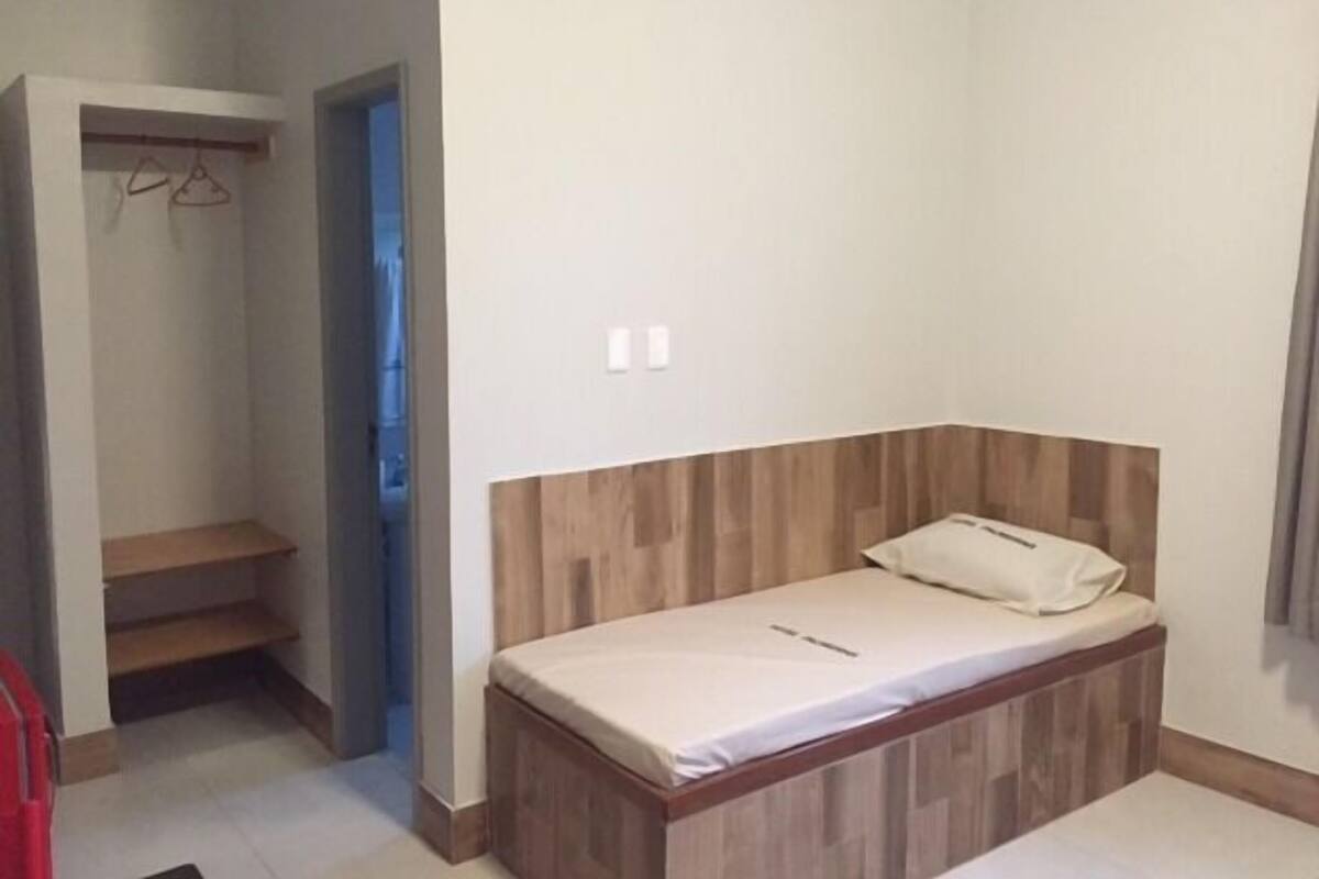 A small, single-bed room with a wooden headboard, a closet with a hanging rack, and a door leading to another room.