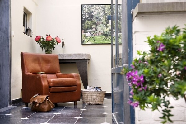 A leather armchair, a wicker basket, and a vase with pink flowers in a hallway with a tiled floor and a framed picture on the wall. - Ganda Rooms & Suites (Ghent)