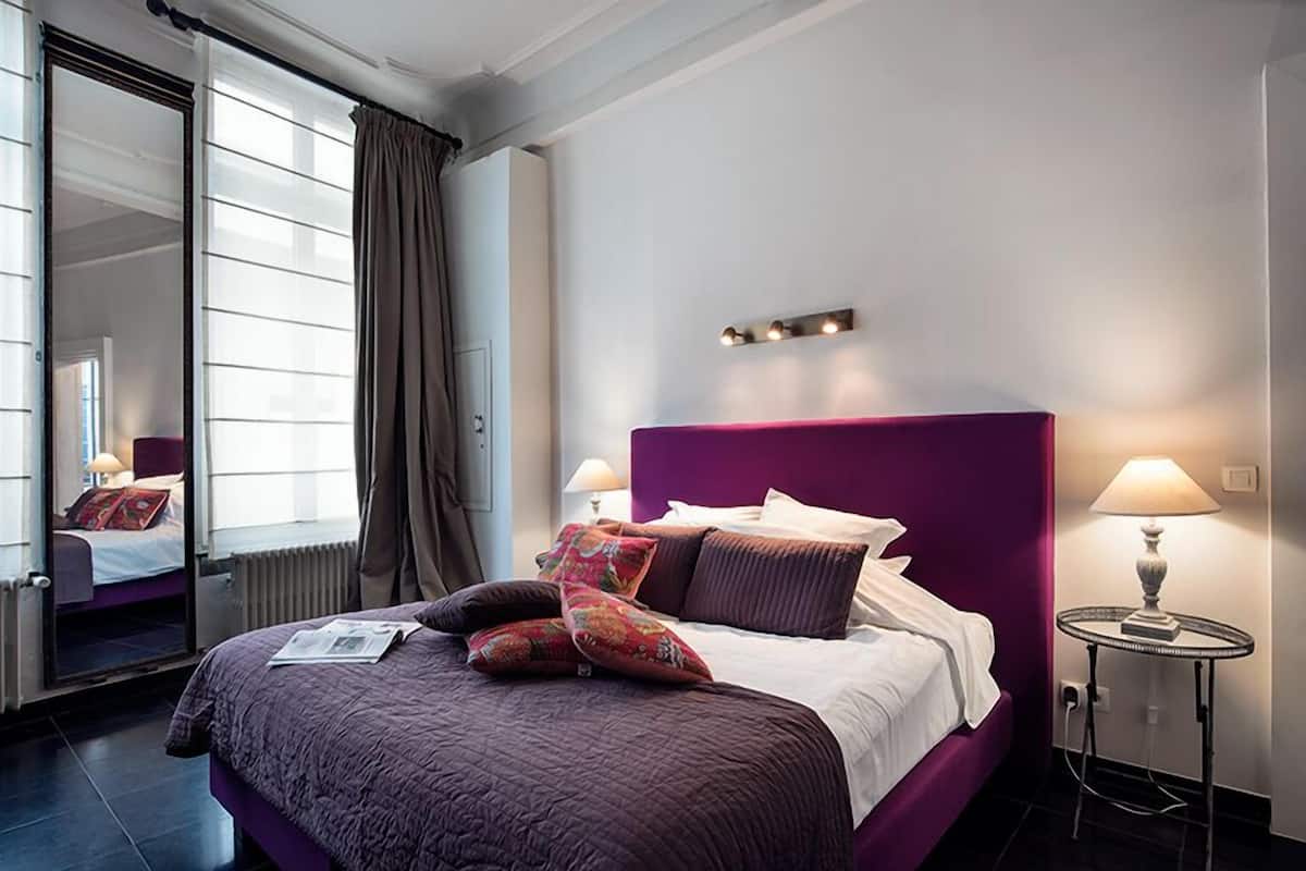 Une chambre d’hôtel moderne avec un grand lit, une literie violette, une table de chevet avec une lampe et une fenêtre avec des rideaux.