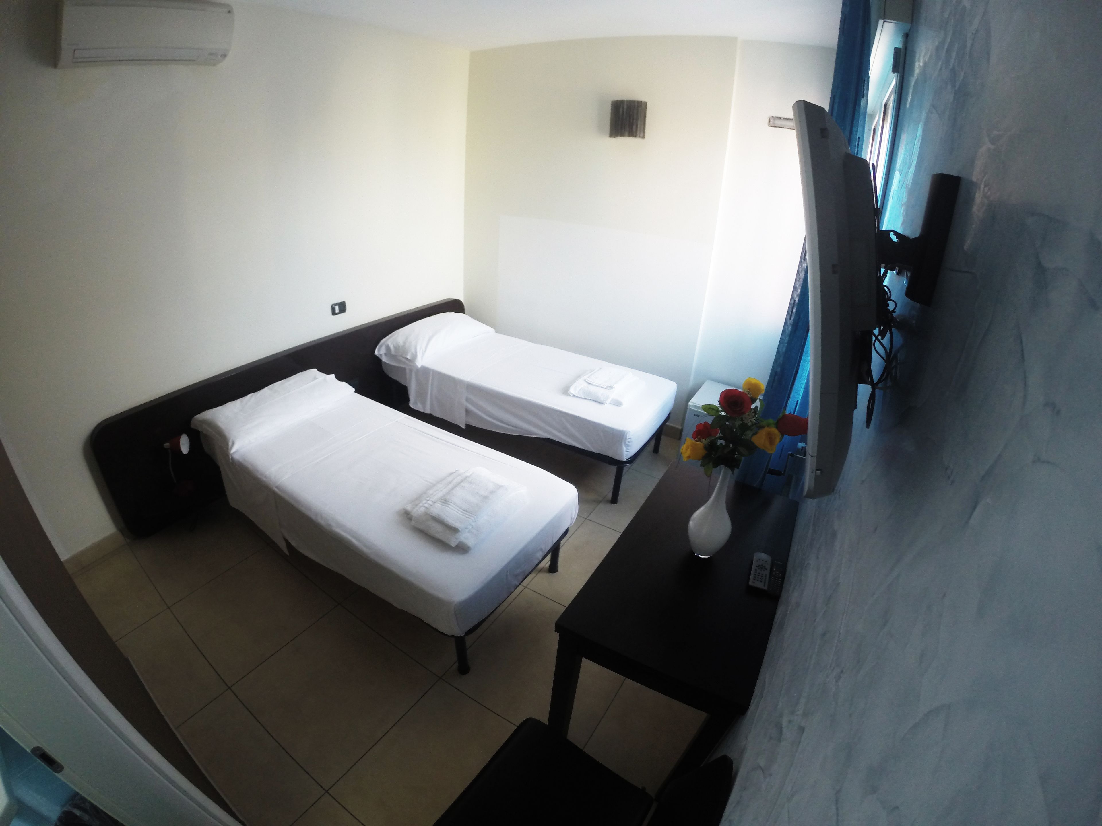 A hotel room with two single beds, a small table with a vase of flowers, and a TV on the wall.