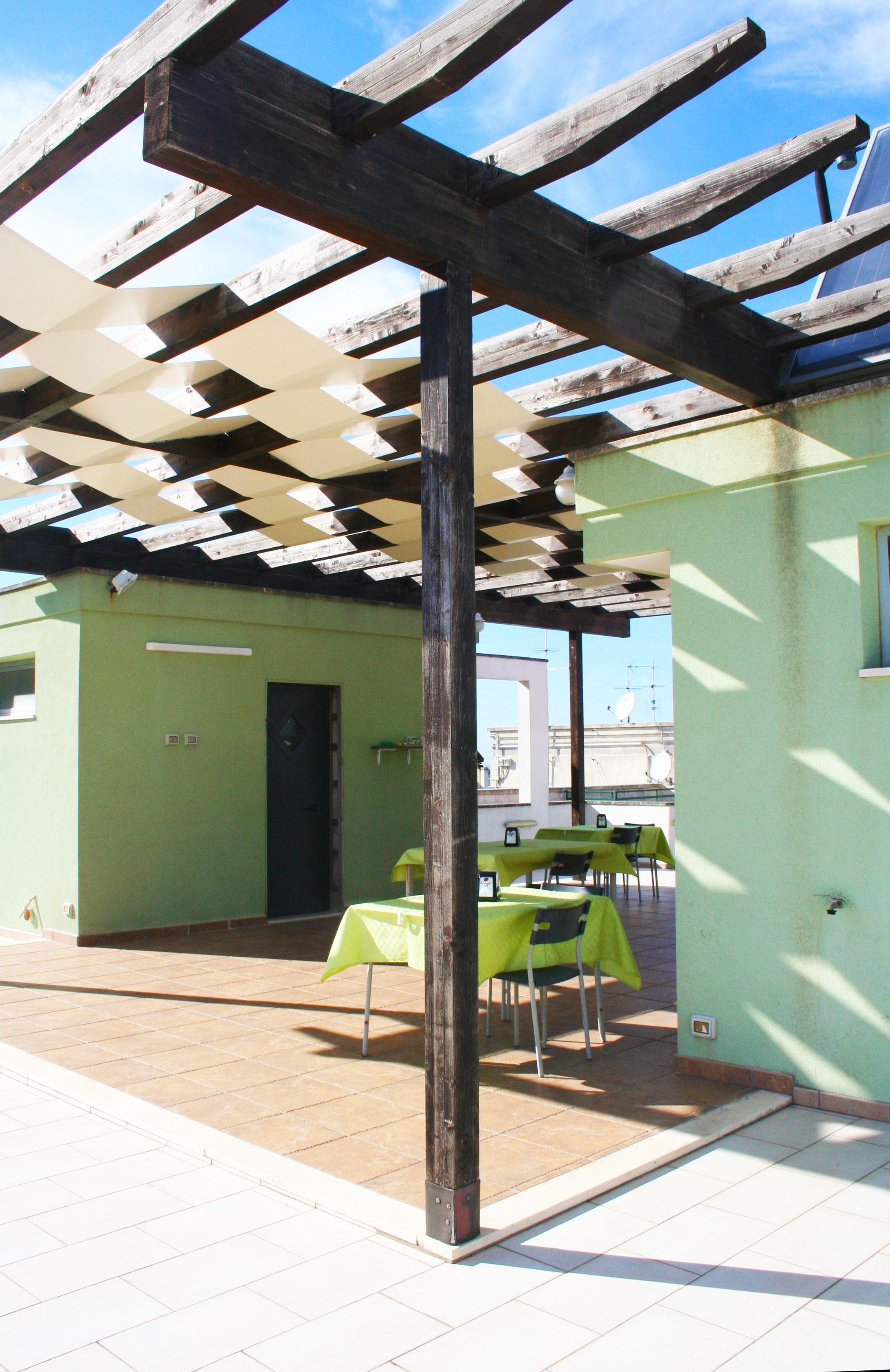 A covered outdoor area with a wooden pergola, green tables, and chairs, suggesting a casual dining or meeting space.