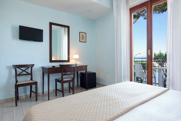 Une chambre d’hôtel comprenant un lit, un bureau, une chaise, un téléviseur et une vue sur la mer, accessible par une porte-fenêtre donnant sur un balcon.