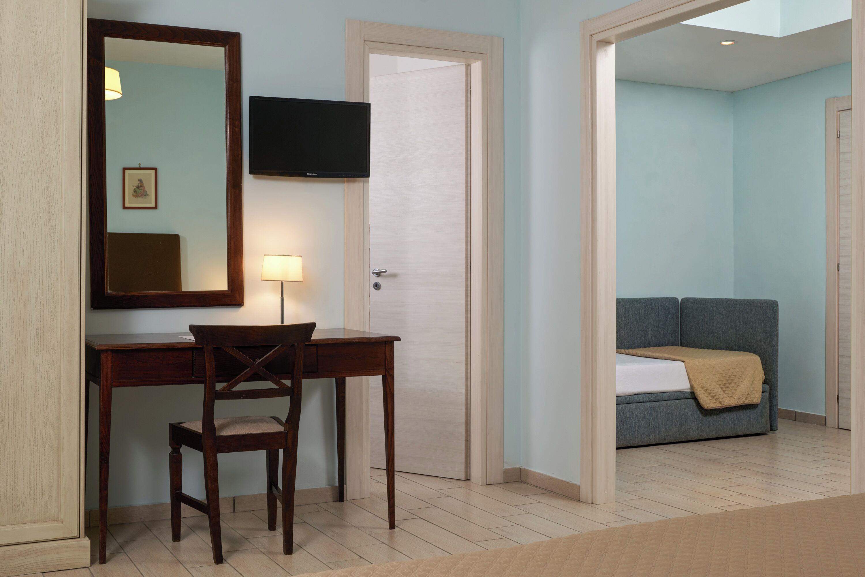 A hotel room with a desk, chair, and a television. There is a bed with a blanket and a sofa in the adjacent room.
