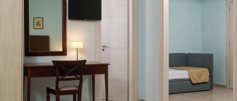 A hotel room with a desk, chair, and a television. There is a bed with a blanket and a sofa in the adjacent room.