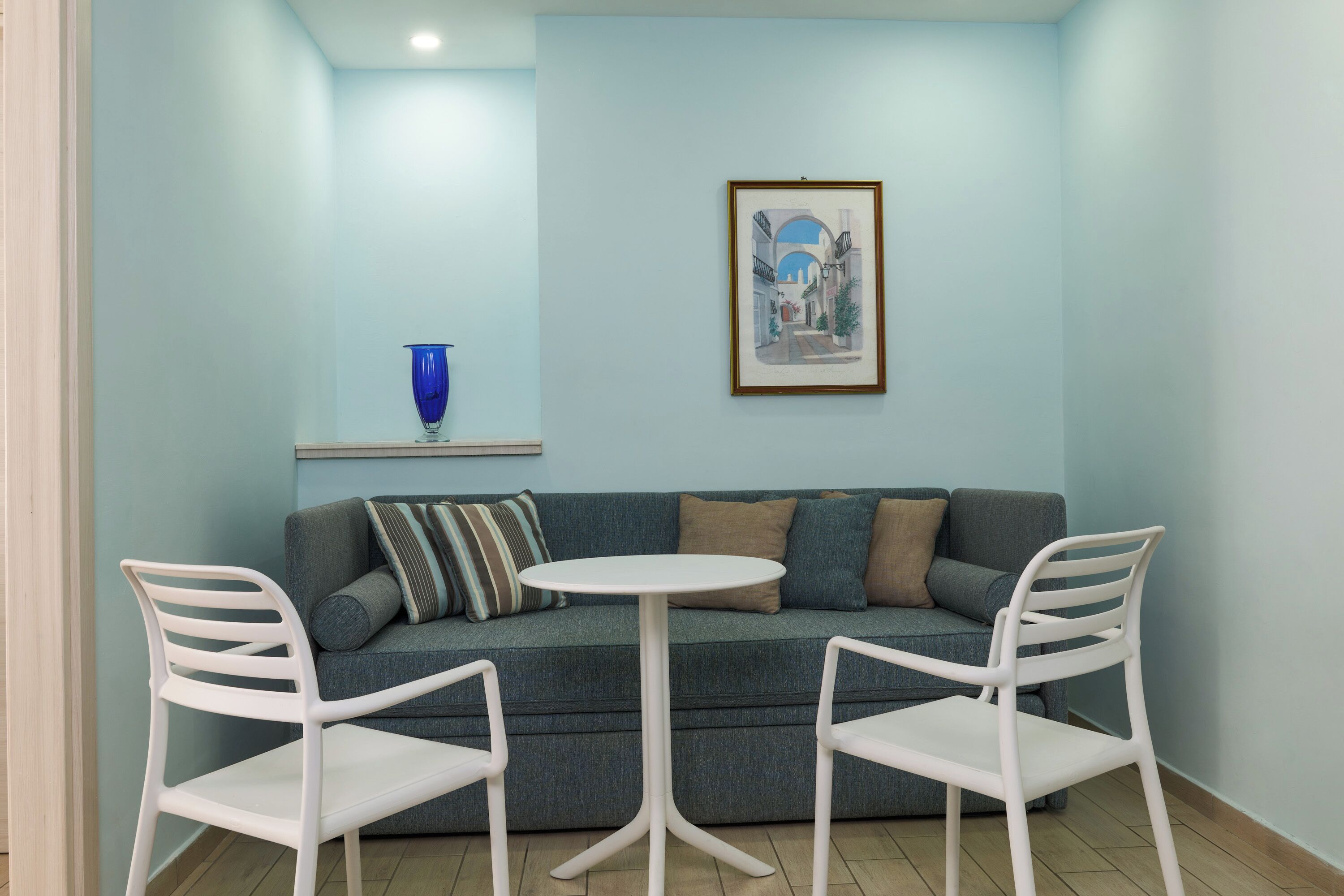 A room with a blue wall, a framed picture, a blue vase, a grey sofa, and two white chairs around a small white table.