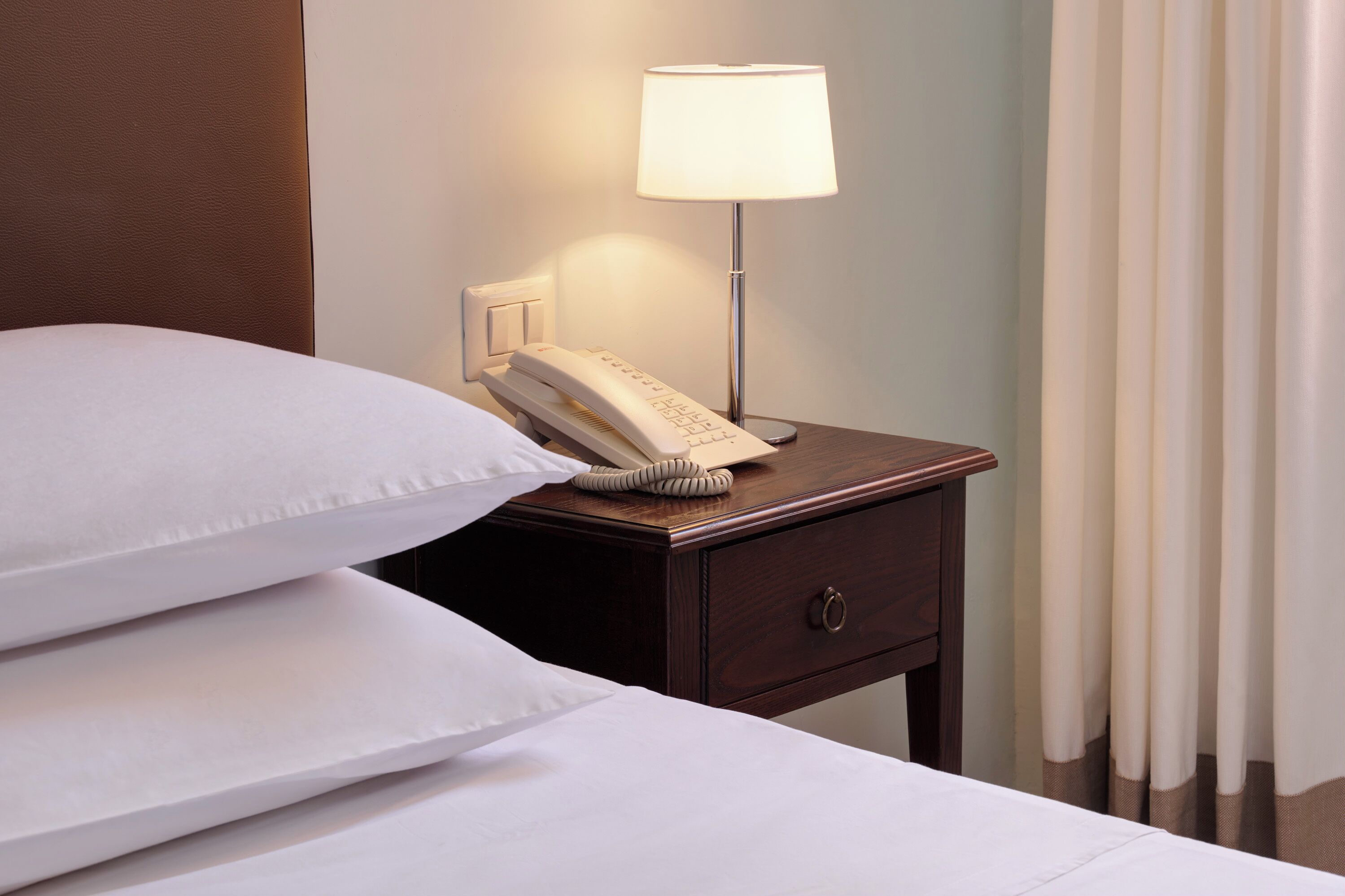 Ein ordentlich bezogenes Bett mit weißen Leinen, ein Nachttischtelefon und eine Lampe auf einem Holznachttisch.