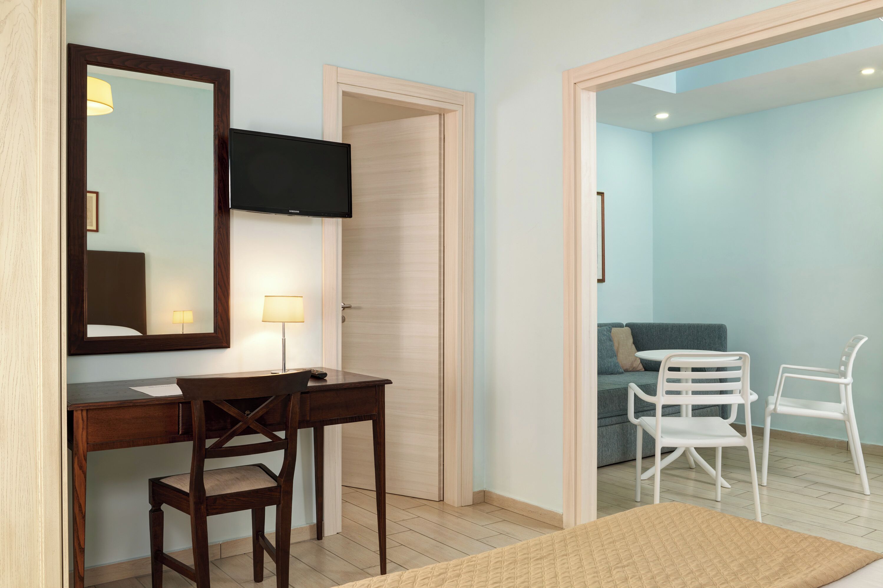 A bedroom with a wooden desk, a chair, a bed, a TV, and a door leading to a living area with a sofa and two chairs.