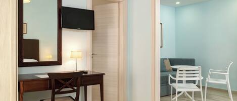 A bedroom with a wooden desk, a chair, a bed, a TV, and a door leading to a living area with a sofa and two chairs.