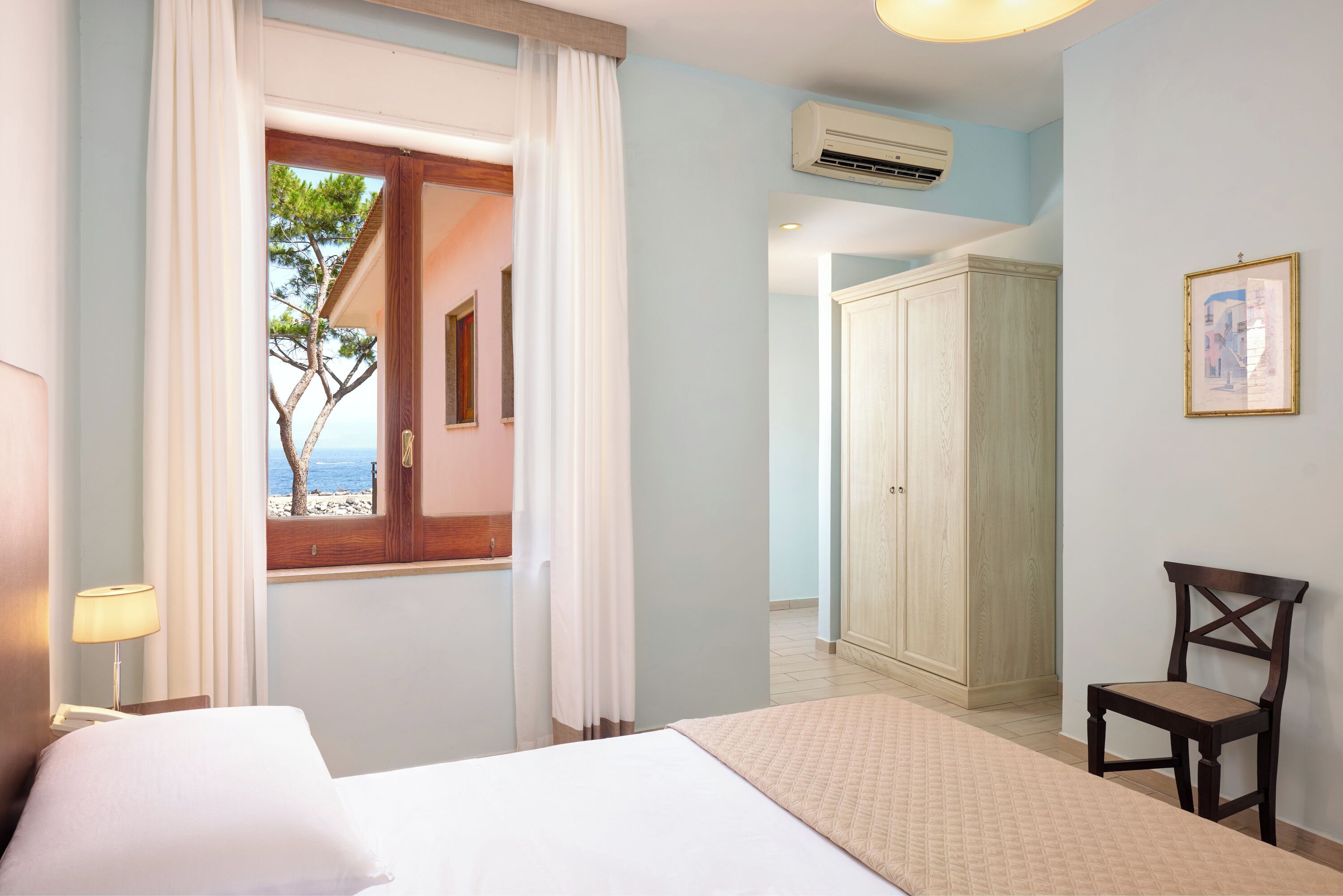 A hotel room with a bed, a chair, a wardrobe, and a window with a view of the sea.