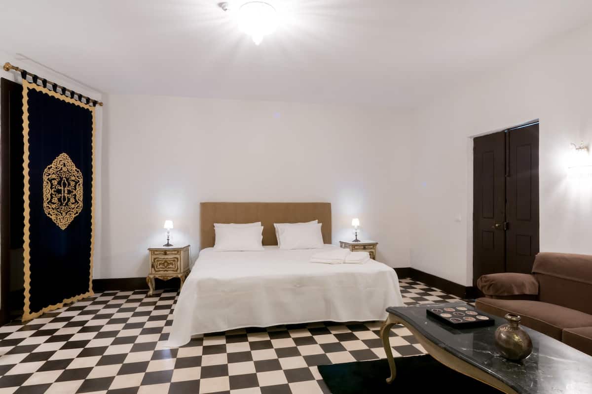 A hotel room with a checkered floor, a bed with white linens, a brown headboard, two bedside lamps, a brown sofa, and a small table with a decorative item.