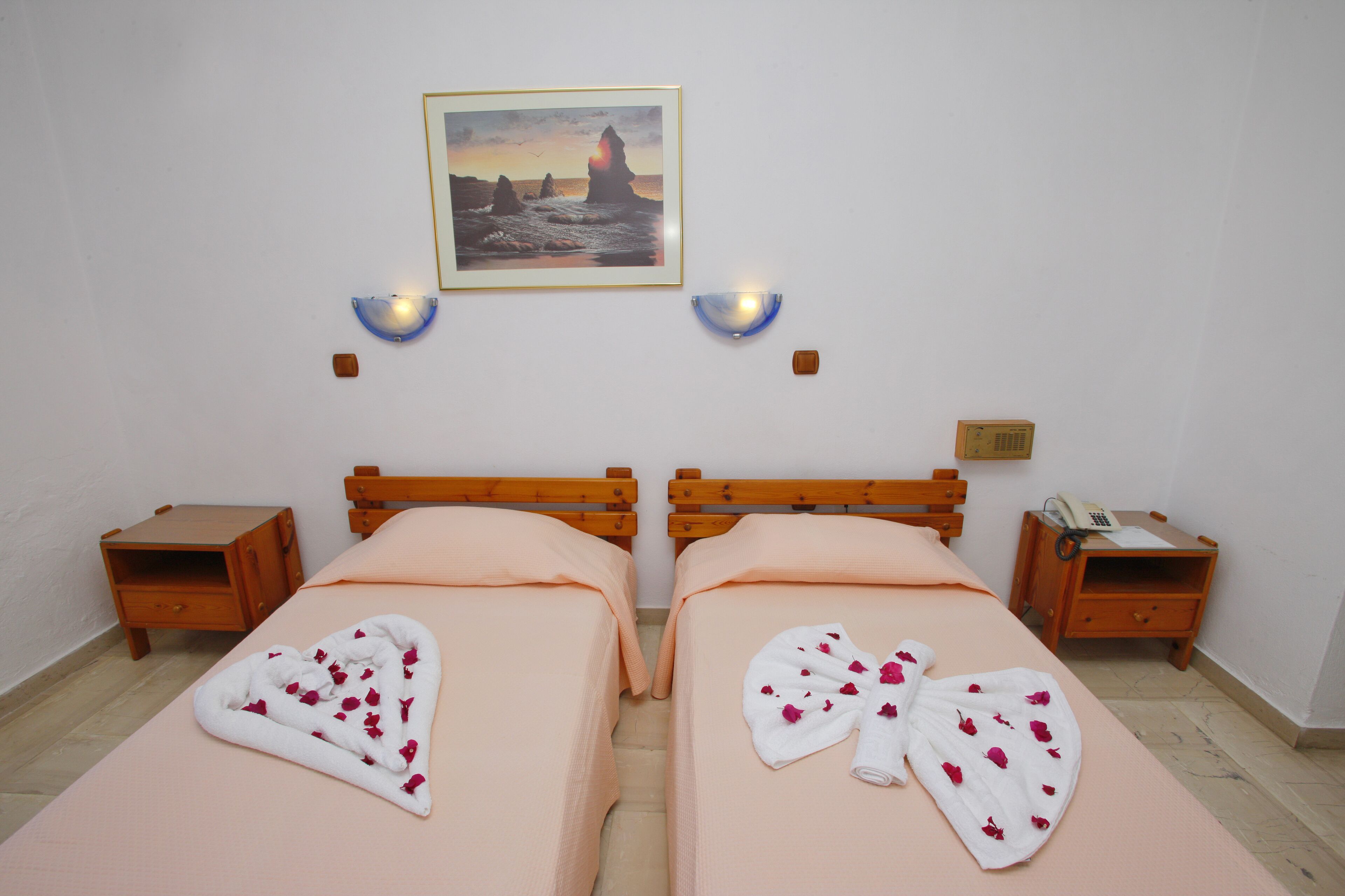 A hotel room with two beds, a painting of a sunset over rocks, and a small table with a phone and papers.